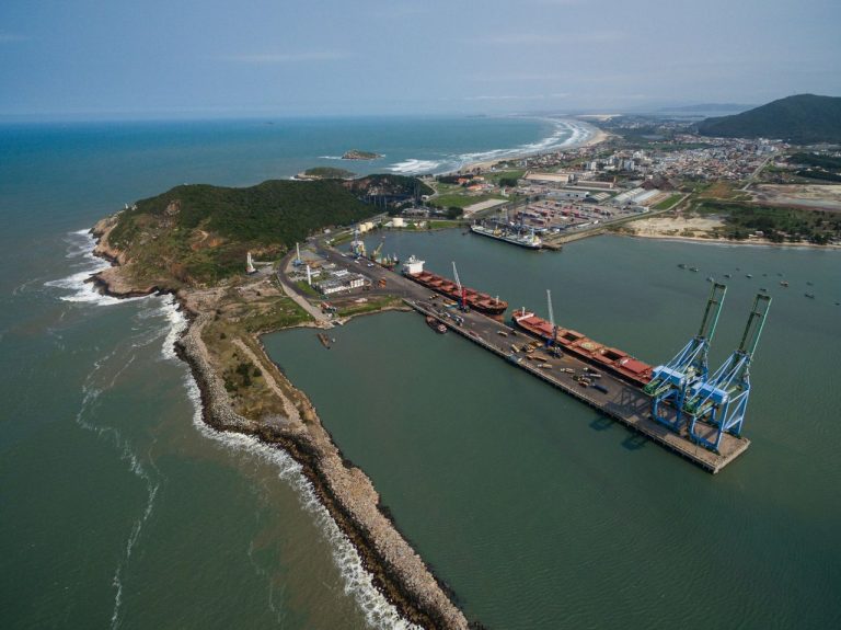 Vista aérea do porto de Imbituba, em Santa Catarina, com cais, guindastes e embarcações junto ao litoral brasileiro.